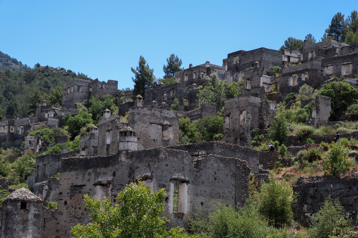 Narrow cobbled lane climbing between ruined roofless stone houses at Kayaköy, pine-covered ridge rising behind